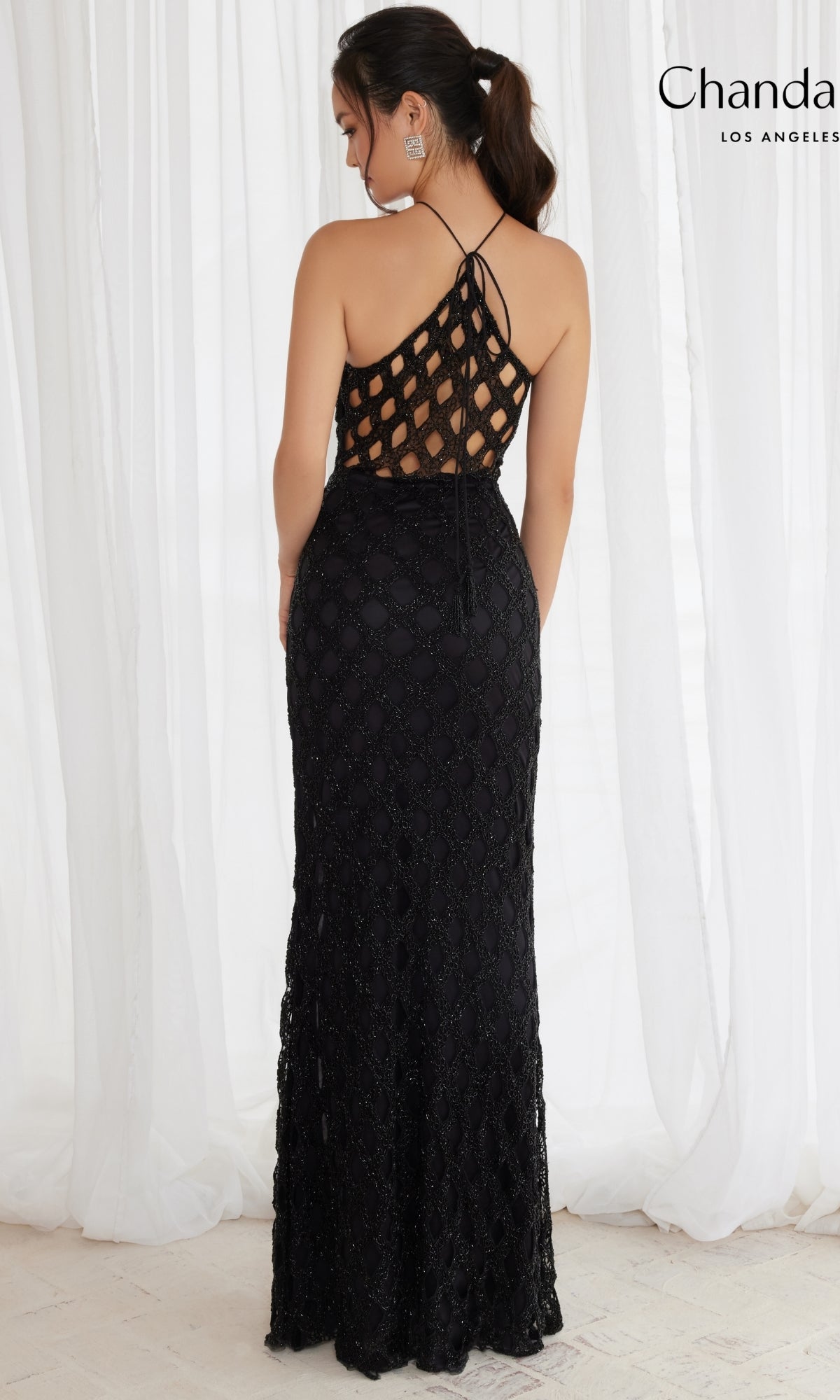 A woman wears the Long Prom Dress 30130 by Chandalier, a floor-length black gown with cut outs and a geometric back design, standing in front of white curtains.